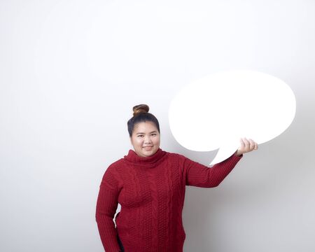 Attractive asian woman in red sweater holding showing blank speech bubble on light gray background,...