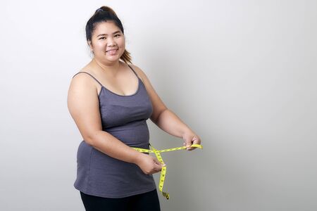 Overweight Asian woman measuring waist before weight loss in studio shot on bright gray background, health problems and overweight, diet conceptの写真素材