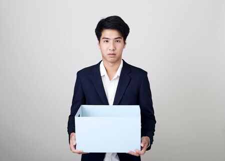Smiling handsome young Thai businessman holding cardboard box of belongings standing studio shot on bright gray background, Financial business conceptsの写真素材