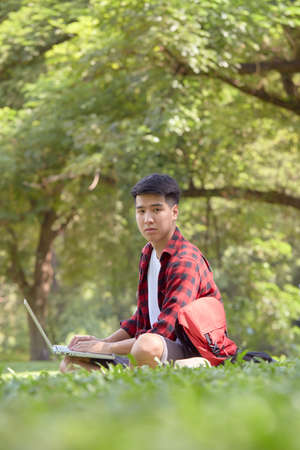Young Asian man backpacker sitting on the grass field on a summer day handsome guy enjoy with outdoor lifestyle on holiday vacationの写真素材