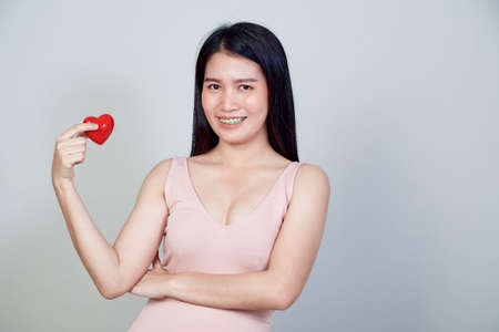 Portrait of beautiful Asian young woman holding pointing red heart shape isolated on light gray...