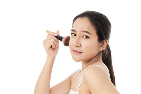Portrait of a beautiful young Asian brunette woman with a clean face. Beauty spa model girl with perfect fresh clean skin applying cosmetic brush. skincare concept. isolated on a white backgroundの写真素材