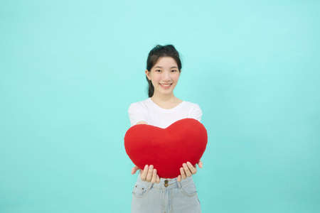Cute cheerful black hair Asian woman smiles with heart pillow shape on light blue studio isolated backgroundの写真素材