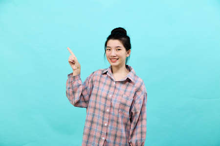 A young Asian woman pretty  pointing looking at ads wearing a plaid shirt standing and feeling happy and confident on blue backgroundの写真素材