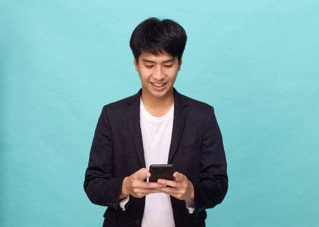 Portrait of a Young smiling handsome Asian man in a semi-formal suit using a mobile phone in light blue isolated studio backgroundの写真素材