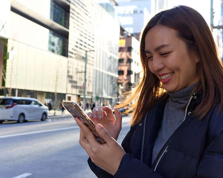 Young happy Asian woman using the mobile app on a smartphone while standing outdoors on road catching a taxi modern cityの写真素材