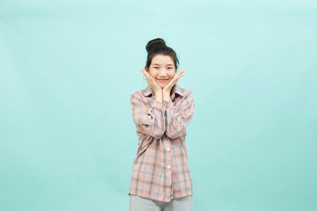A pretty female model with a ginger hair knot has a warm pleasant smile, keeps their hand under their chin, wearing a shirt, and jeans on bright blue copy space. Happiness conceptの写真素材