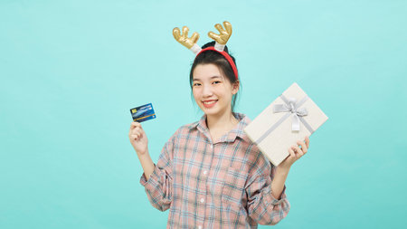 Cheerful cute Asian woman holding a gift box with a credit card on bright blue color background. Christmas and new year sales joy conceptの写真素材