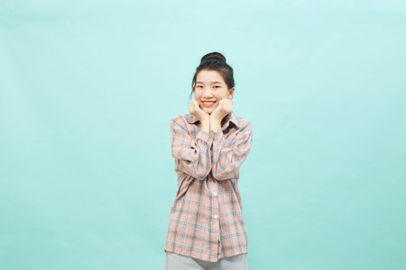 A pretty female model with a ginger hair knot has a warm pleasant smile, keeps their hand under their chin, wearing a shirt, and jeans on bright blue copy space. Happiness conceptの写真素材