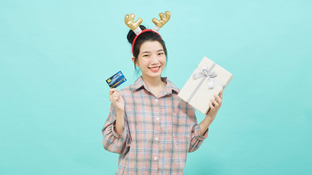 Cheerful cute Asian woman holding a gift box with a credit card on bright blue color background. Christmas and new year sales joy conceptの写真素材