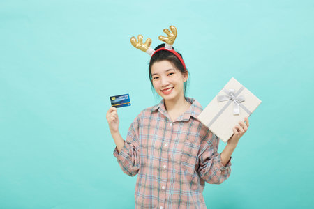 Cheerful cute Asian woman holding a gift box with a credit card on bright blue color background. Christmas and new year sales joy conceptの写真素材