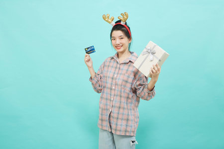 Cheerful cute Asian woman holding a gift box with a credit card on bright blue color background. Christmas and new year sales joy conceptの写真素材