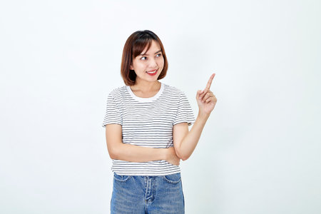 Portrait of smiling attractive Asian female in a casual t-shirt and blue jeans pointing finger upper white background. Copy spaceの写真素材