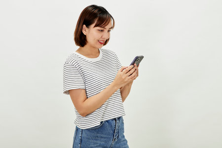 Smiling Asian woman using a smartphone, standing against texting on a mobile phone on white background, people and technology conceptの写真素材