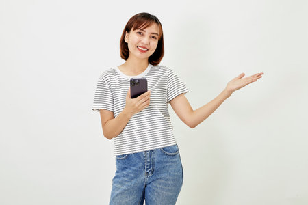 Young Asia woman holding showing mobile phone and pointing a finger empty copy space, showing promo text, standing with a smartphone against on white backgroundの写真素材