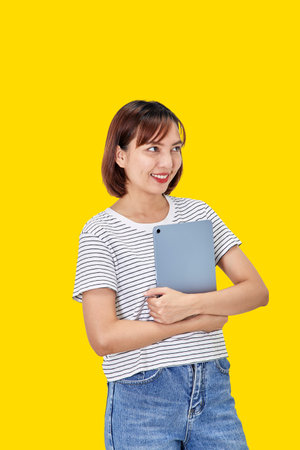 Portrait of a smiling beautiful Asian woman freelancer using a digital tablet and looking pleased with the device screen, shopping online, on yellow backgroundの写真素材