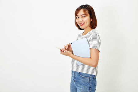 Charming young Asian female employee with short brown hair wearing t-shirt and jeans smiling and holding a digital tablet with smartphone standing on white background copy spaceの写真素材
