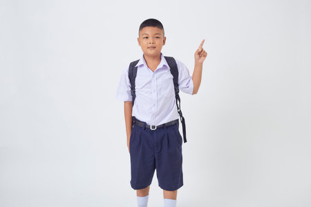 A young Asian cute boy standing in a Thai school uniform with a backpack bag and book on a white background banner. Back to schoolの写真素材