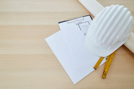Construction tools and a white hard hat with a measuring stick, paper house building plan on a wooden work table copy space for textの写真素材