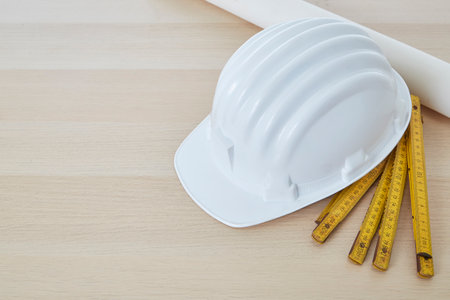 Construction tools and a white hard hat with a measuring stick, paper house building plan on a wooden work table copy space for textの写真素材