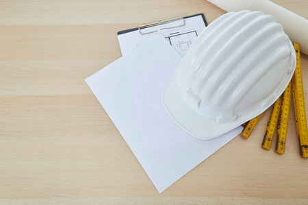 Construction tools and a white hard hat with a measuring stick, paper house building plan on a wooden work table copy space for textの写真素材