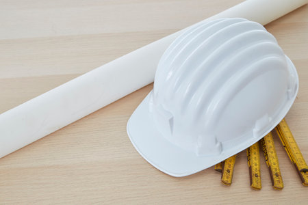 Construction tools and a white hard hat with a measuring stick, paper house building plan on a wooden work table copy space for textの写真素材