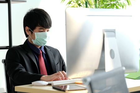 Man in the office wear mask to prevent corona virus. He looks to the PC and see information on virus. On the table have tablet, keyboard and mouse. Protection virus and business concept.の写真素材