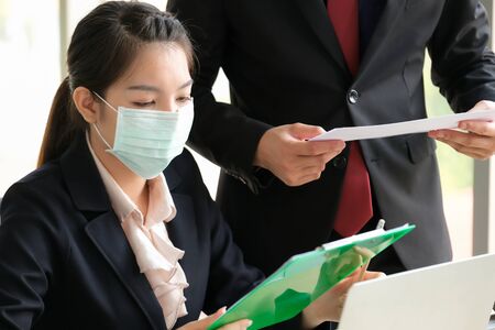 Women in the office wear mask to prevent corona virus. She looks at clipboard and talk with boss on the corona virus situation. Feeling very sad. Protection virus and business concept.の写真素材