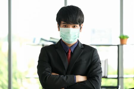 Man in the office wearing mask to prevent corona virus. He is standing in front of the desk and folded his arms with blurred background. He wear the black suit. Protection virus and business concept.の写真素材