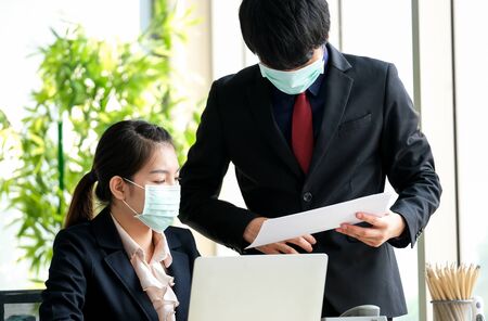 Women in the office wear mask to prevent corona virus. She is listening her boss to inform on the progress of work. Notebook and telephone on the desk. Protection virus and business concept.の写真素材