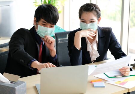 Man and woman in the office wear mask to prevent corona virus. They see information in the computer. There are notebook pencil and paper on the table. Protection virus and business concept.の写真素材