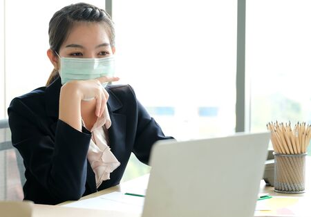 Woman in the office wear mask to prevent corona virus. Close her mouth using hand.  She looks to the laptop and see information on virus. Blurred background with sunlight. Protection virus concept.の写真素材