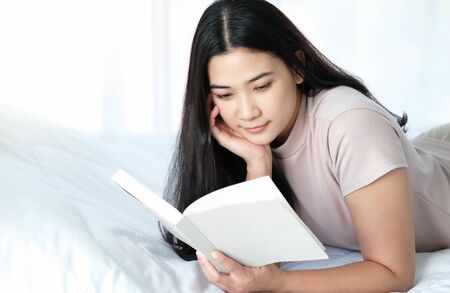 Pretty Asian woman laying down on the white bed and reading a book with copy space under sunlight. Feeling happy, smile and relaxation. Working at home and holiday concept.の写真素材