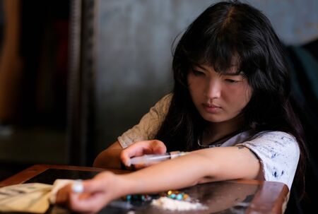 Young Asia woman sitting in the abandoned house and feeling hopeless. There are many drug and gun on the table.  A girls used syringe to input drug at her arm. Drug addict and hopeless conceptの写真素材