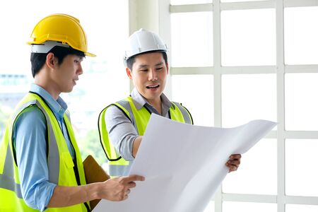 Two engineers man standing in front of the site. Left engineer holding blueprint diagram and right engineer point his hand to  drawing diagram in blueprint. Engineering and construction concept.の写真素材