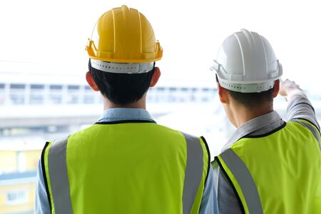 Two engineers man standing in front of the site. Right engineer holding paper roll and point to construction area and explain on progress to other engineer. Engineering and construction concept.の写真素材