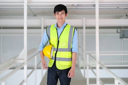 Junior engineer standing in the plant holding helmet with copy space. Wearing safety vest. Feeling happy and smile on work. Engineering and design concept.の写真素材