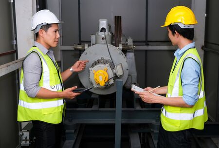 Senior engineer point to the lift motor and explain to junior engineer. Feeling fun on work at the control room of plant. Wearing safety vest and safety helmet. Engineering and control room concept.の写真素材