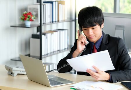 Young businessman is talking on phone with customer. He is working with laptop on the table in the modern office and behind have bookshelf with blurred background. Business and marketing concept.の写真素材