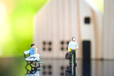 Miniature people : A man who wear mask to protect pollution, corona virus (COVID-19) sitting on the Bench. Traveler standing in front of house. Healthcare and Social distancing concept.の写真素材