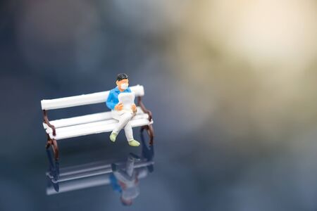 Miniature people : A man who wear mask to protect pollution, corona virus (COVID-19) sitting on the bench and read newspaper under sunlight with blurred background. Healthcare and relaxation concept.の写真素材