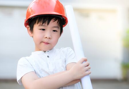 Young Asia boy in gray T shirt wearing a orange safety helmet and holding blueprints in the garden at home under sunlight with blurred background.  Engineering and learning education concept.の写真素材