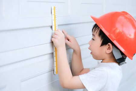 Young Asia boy in gray T shirt wearing a orange safety helmet and used triangular reduction scales to measure the door in the garden at home. Engineering and learning education concept.の写真素材