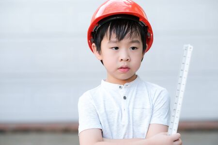 Young Asia boy in gray T shirt wearing a orange safety helmet and holding triangular reduction scales in the garden at home. Feeling happy, fun and smile. Engineering and learning education concept.の写真素材