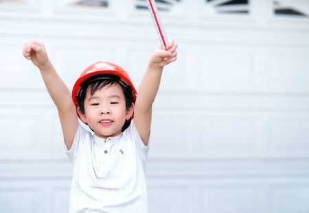 Young Asia boy in gray T shirt wearing a orange safety helmet and holding triangular reduction scales and raise hand up in the garden at home. Feeling happy and fun. Engineering and education concept.の写真素材