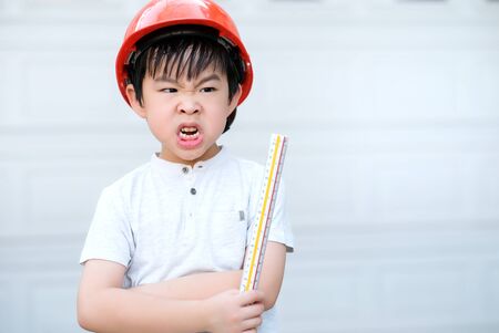 Young Asia boy in gray T shirt wearing a orange safety helmet and holding triangular reduction scales in the garden at home. Feeling unhappy. Engineering and learning education concept.の写真素材