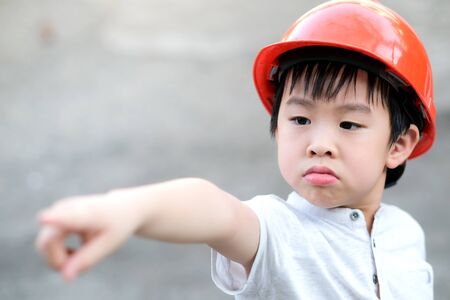 Young Asia boy in gray T shirt wearing a orange safety helmet stand in the garden at home under sunlight. Feeling angry and unhappy. Point his hand out. Engineering and learning education concept.の写真素材