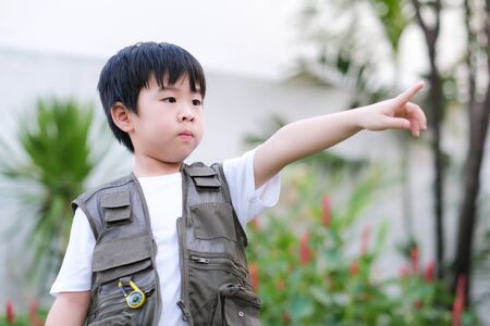 Curious little young Asian boy standing in the garden and point finger out. Exploring the world, outdoors activity. Feeling fun and happy. Boy wear green outdoors vest. Holiday, education conceptの写真素材