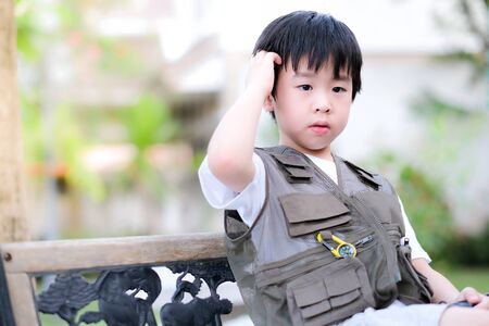 Young Asian boy sitting on the chair in the garden. Exploring the world, outdoor activity. Feeling confuse and lost direction. Boy wear green outdoors vest and scratch head. Holiday, learning conceptの写真素材