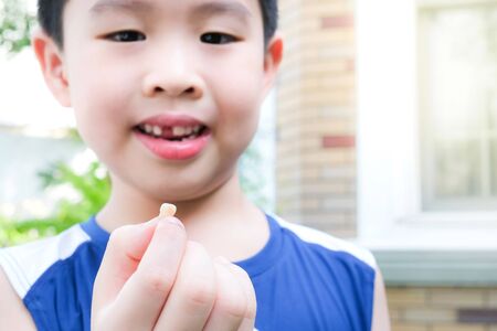 Young  Asia boy (6 years old) surprised to lost his top front milk teeth. He hold a teeth on his finger in the garden under sunlight with copy space. Feeling happy and smile. Healthcare concept.の写真素材